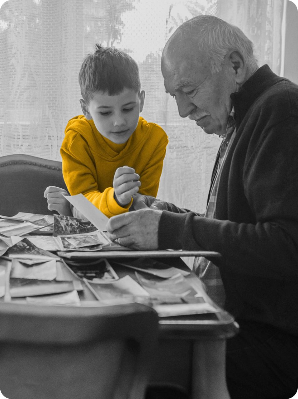 Elderly man looking at pictures with child in yellow shirt sitting at a dining table. Black and white photo only yellow shirt is in color.