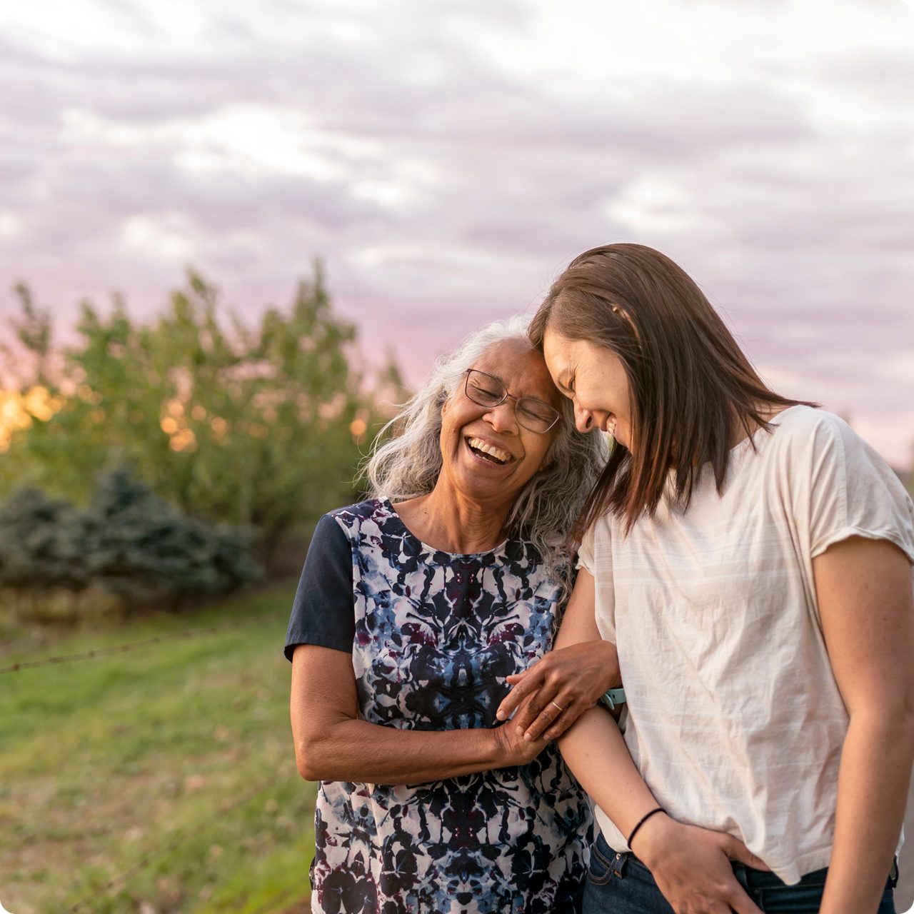 Two smiling women standing side-by-side outdoors, locking arms and enjoying a joyful moment in nature.