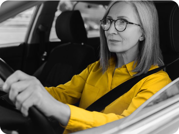 Woman with glasses wearing a yellow shirt driving a car. Black and white photo only yellow shirt is in color. 