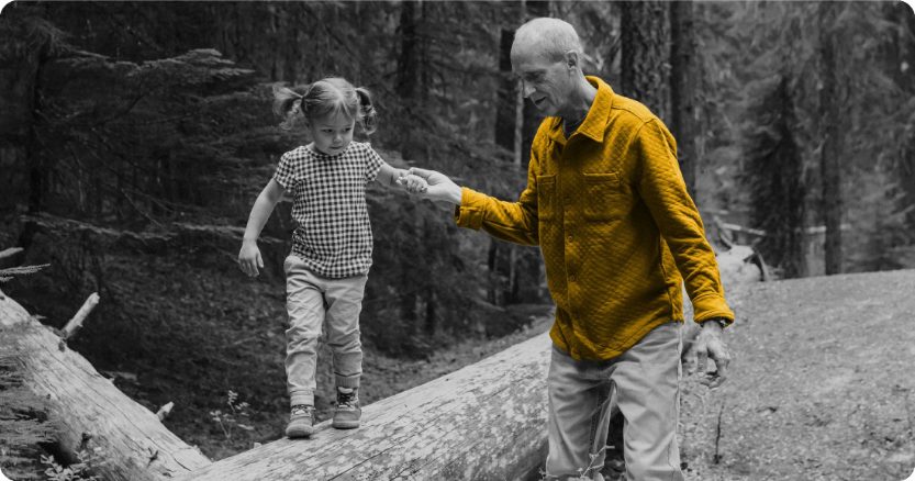 Man in yellow shirt assisting grandchild walking on a big fallen tree trunk. Black and white photo only yellow shirt is in color. 