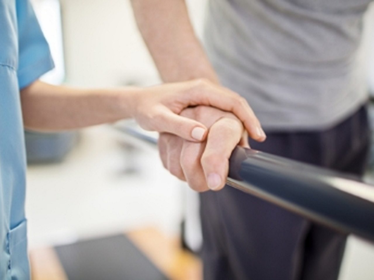 Ein Mitarbeiter des Gesundheitswesens stützt die Hand einer Person, die sich an einer Parallelstange festhält, möglicherweise während einer Physiotherapie- oder Rehabilitationsmaßnahme.