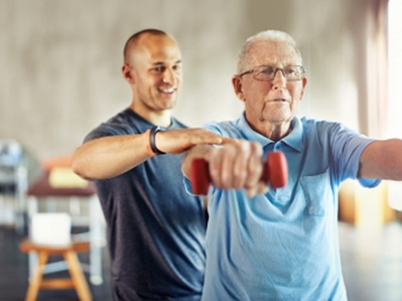 Ein jüngerer Mann hilft einem älteren Mann beim Heben kleiner Hanteln, beide stehen in einem Fitnessstudio oder einer Fitnesseinrichtung.