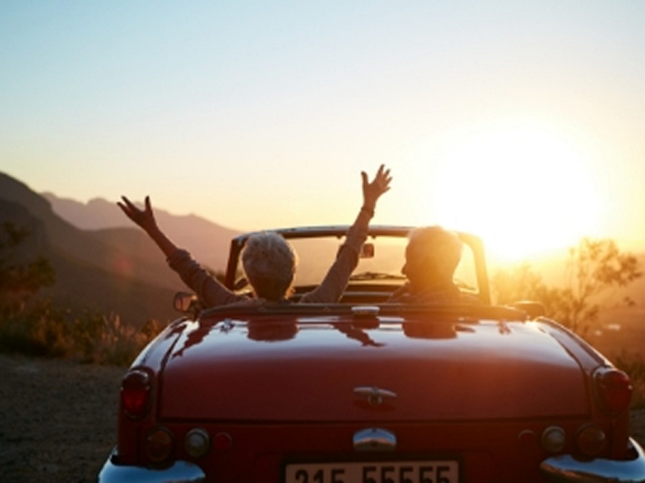 Zwei Personen in einem roten Cabrio mit offenem Verdeck heben ihre Arme, während sie in einer malerischen Berglandschaft dem Sonnenuntergang entgegenfahren.