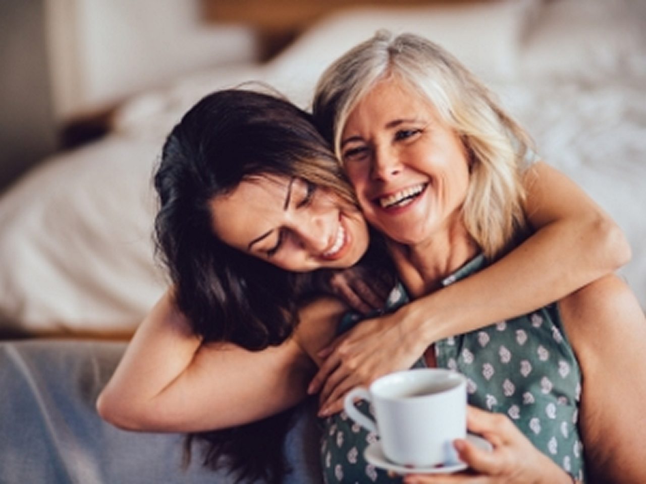 Zwei Frauen sitzen lächelnd und sich umarmend in einem Haus, eine hält eine weiße Kaffeetasse.