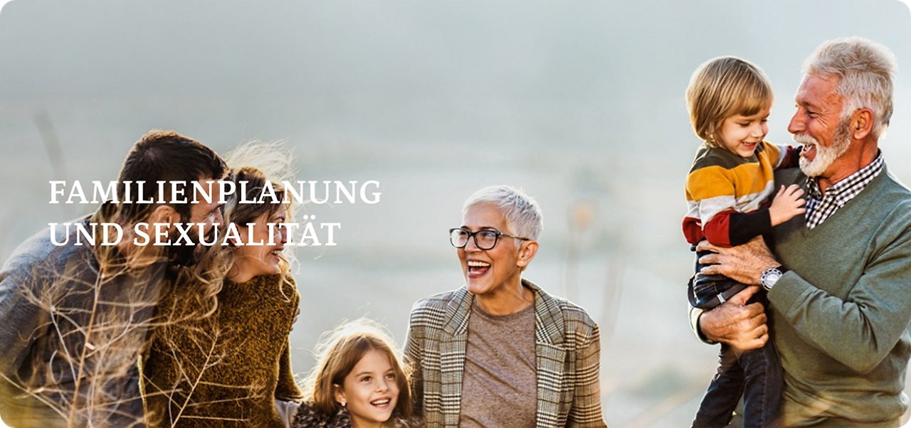 Drei Erwachsene, zwei Kinder und ein älterer Mann stehen gemeinsam auf einem Feld, lächeln und lachen, während im Hintergrund die Berge zu sehen sind.