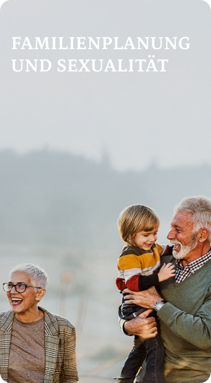 Drei Erwachsene, zwei Kinder und ein älterer Mann stehen gemeinsam auf einem Feld, lächeln und lachen, während im Hintergrund die Berge zu sehen sind.