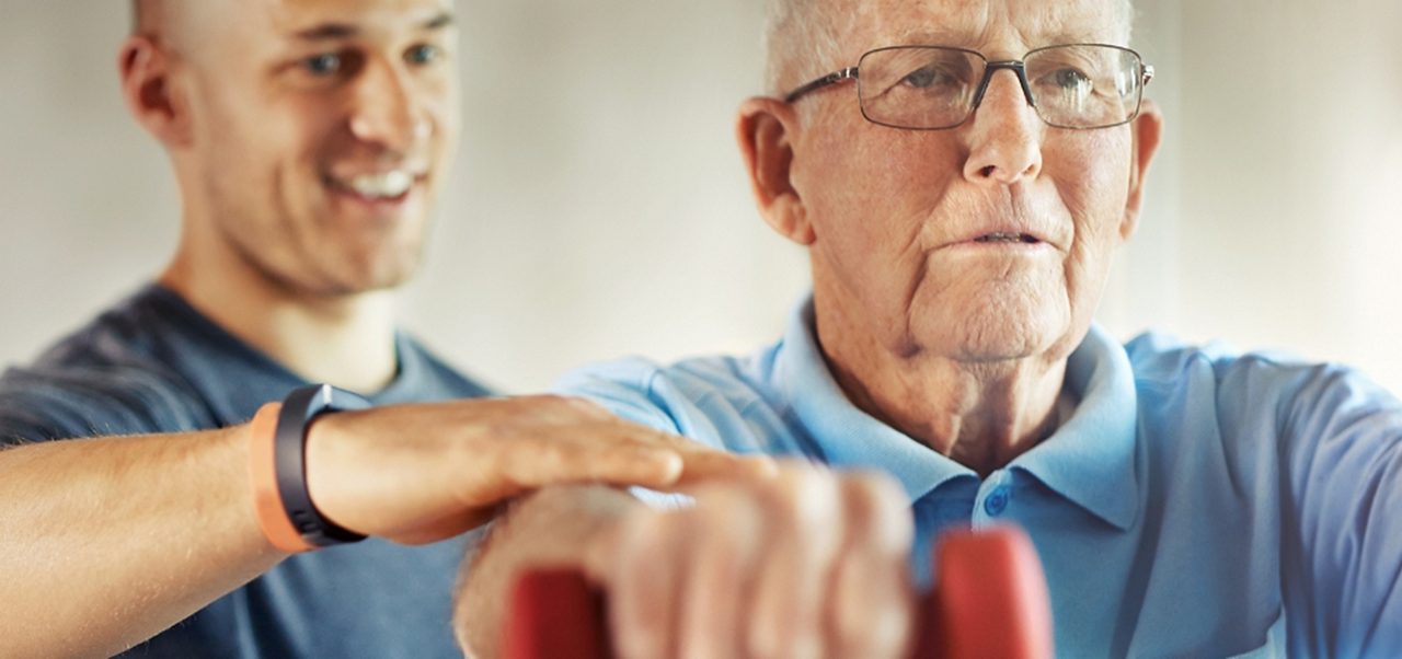 Ein jüngerer Mann hilft einem älteren Mann beim Heben kleiner Hanteln, beide stehen in einem Fitnessstudio oder einer Fitnesseinrichtung.