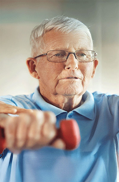 Ein jüngerer Mann hilft einem älteren Mann beim Heben kleiner Hanteln, beide stehen in einem Fitnessstudio oder einer Fitnesseinrichtung.