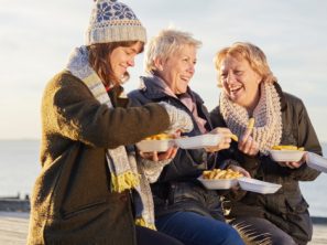 Drei Frauen, die Mützen und Schals anhaben und lachen, während sie Pommes essen