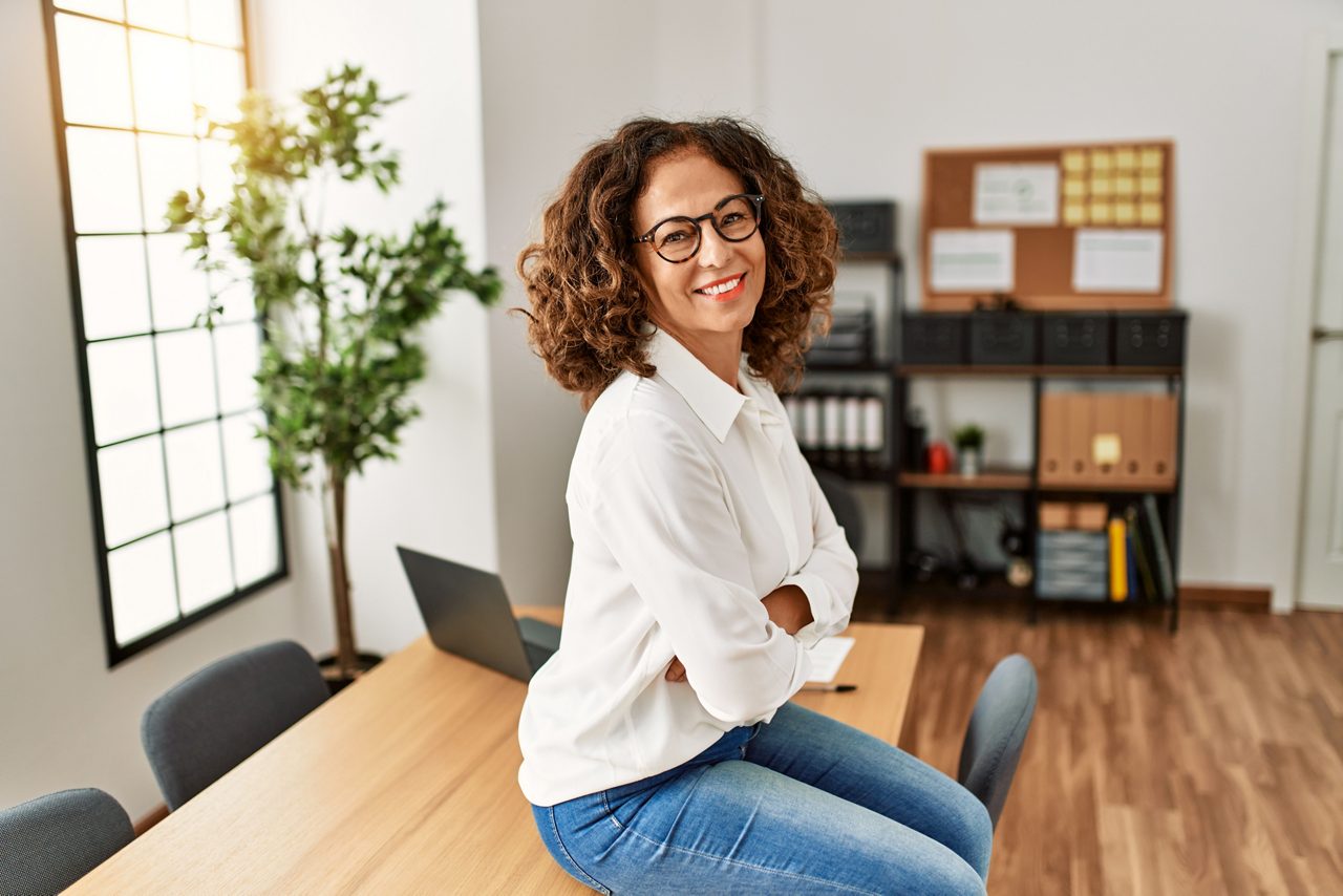  Donna ispanica di mezza età in menopausa, con braccia conserte e seduta su un tavolo, si gira per guardare sorridente e fiduciosa nella telecamera.