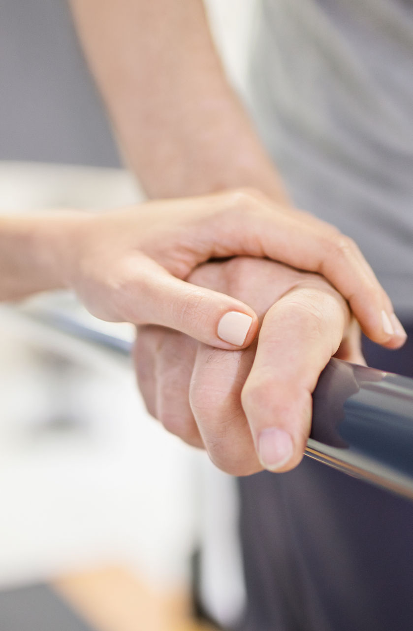 Ein Mitarbeiter des Gesundheitswesens stützt die Hand einer Person, die sich an einer Parallelstange festhält, möglicherweise während einer Physiotherapie- oder Rehabilitationsmaßnahme.