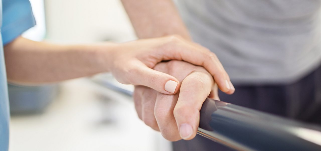 Ein Mitarbeiter des Gesundheitswesens stützt die Hand einer Person, die sich an einer Parallelstange festhält, möglicherweise während einer Physiotherapie- oder Rehabilitationsmaßnahme.