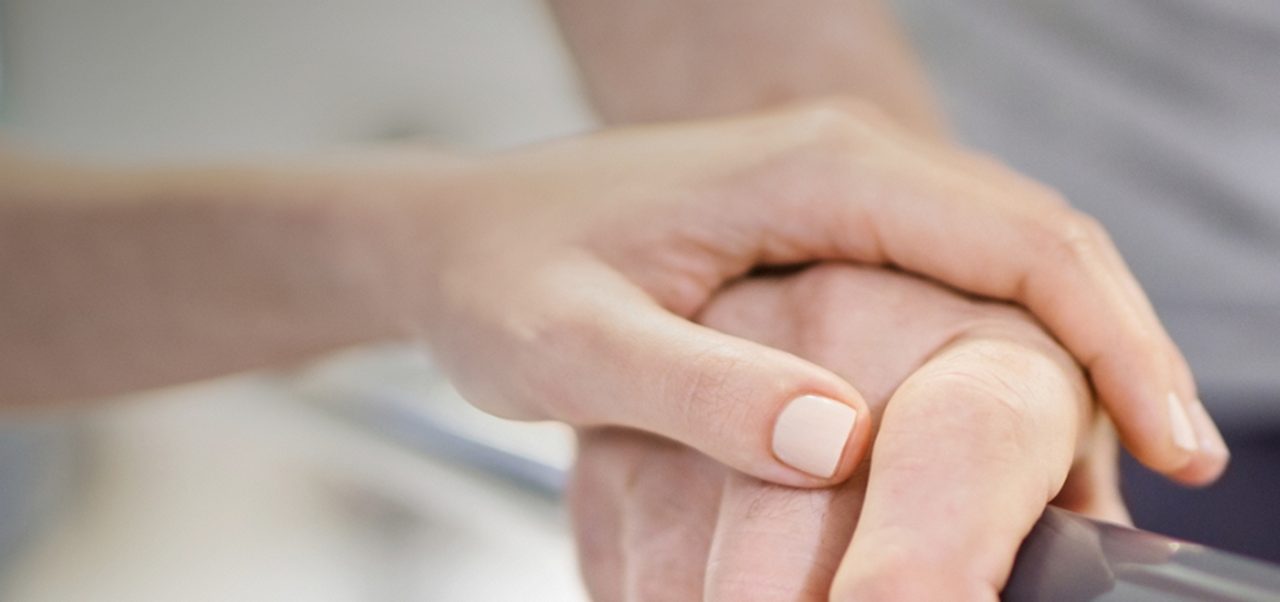 Ein Mitarbeiter des Gesundheitswesens stützt die Hand einer Person, die sich an einer Parallelstange festhält, möglicherweise während einer Physiotherapie- oder Rehabilitationsmaßnahme.