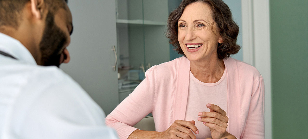 Happy older mature female patient talking to indian male doctor at appointment. Healthy smiling senior old woman feeling cheerful about good news at medical consultation. Elderly health medical care.