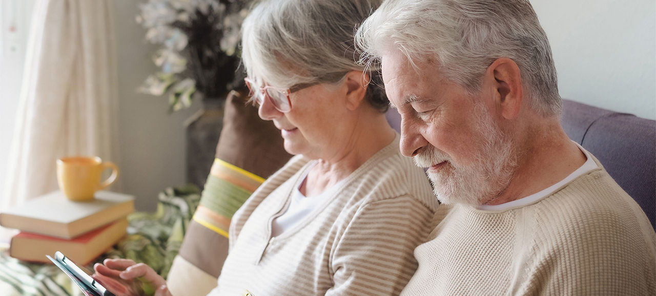 Relaxed senior couple sitting together on home sofa enjoying technological devices, woman browsing on digital tablet, man using phone