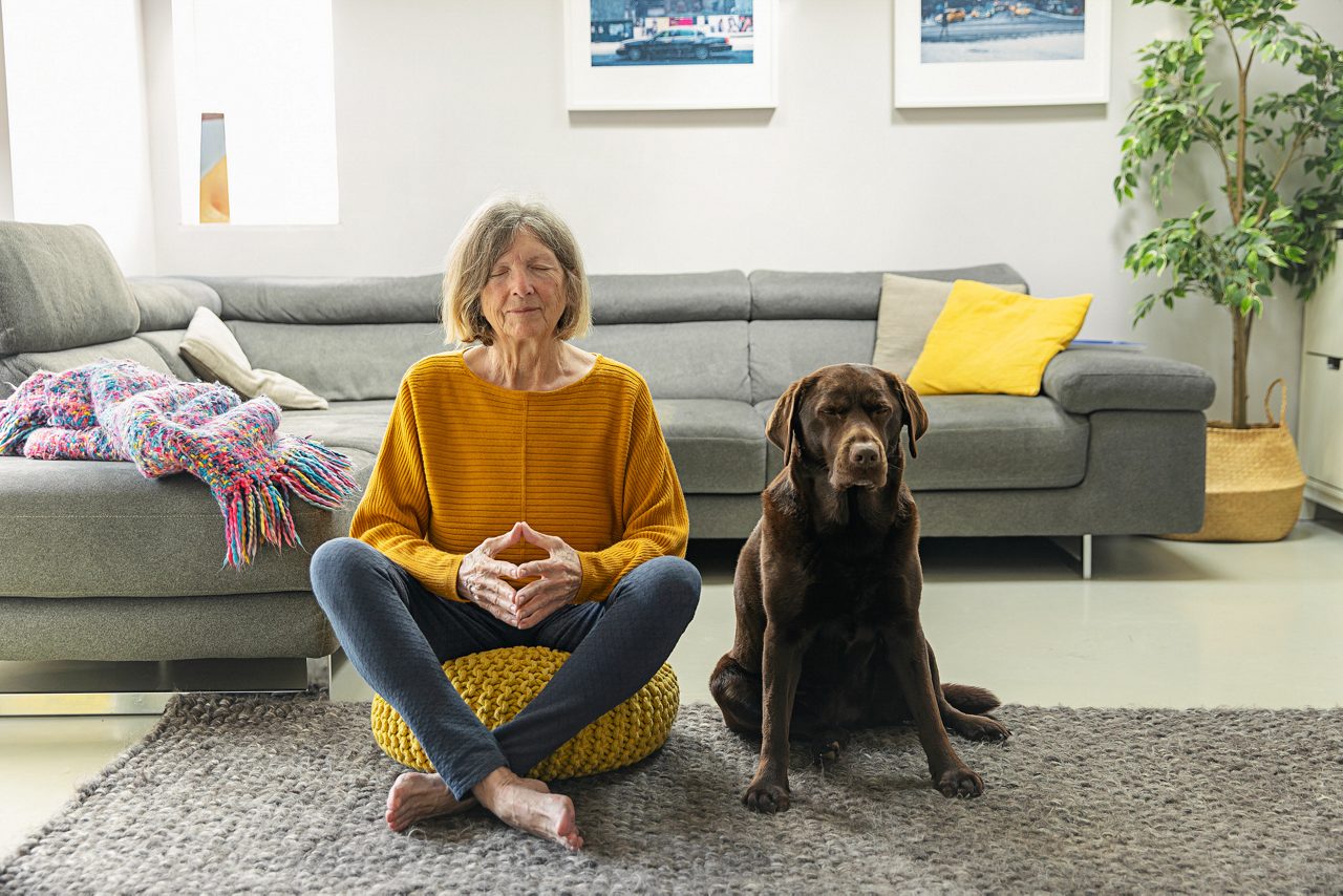 A senior woman (in her 70s who is undergoing targeted treatment for Stage 4 non small-cell lung cancer (NSCLC)) meditates in her living room with her chocolate labrador dog. Their faces displays their positivity.