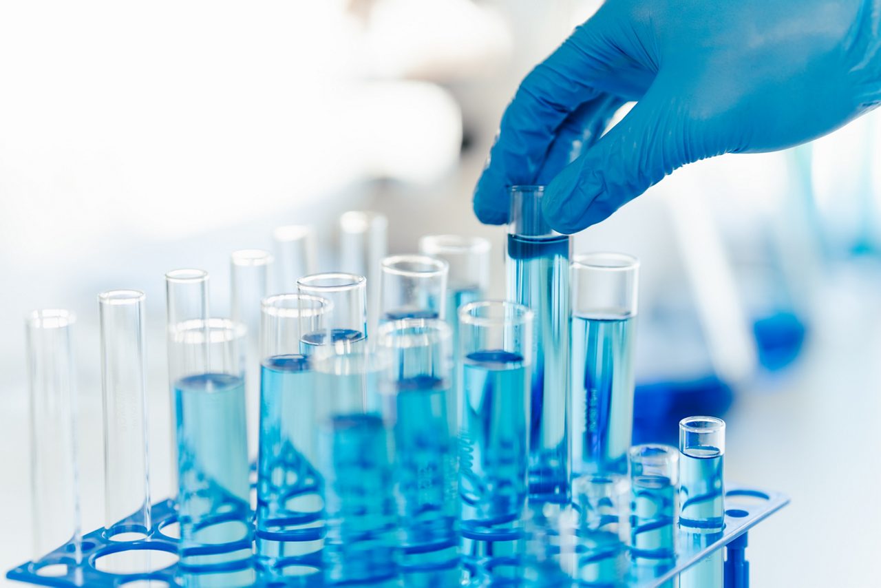 眼科領域の研究を推進するアステラスの最先端研究所で、手袋をした研究者の手が化学物質入りの試験管を持ち上げる様子が描かれています。