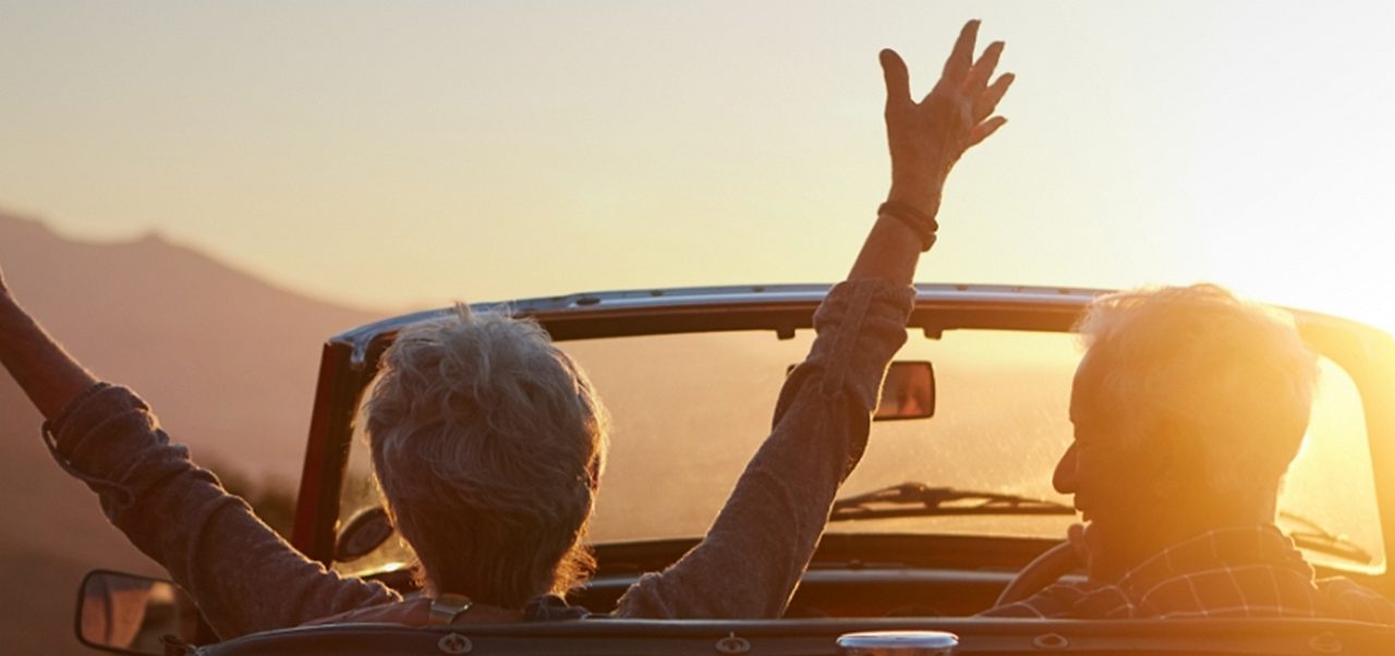 Zwei Personen sitzen in einem roten Cabrio mit offenem Verdeck und erhobenen Armen und blicken auf einen Sonnenuntergang über einer Berglandschaft. 