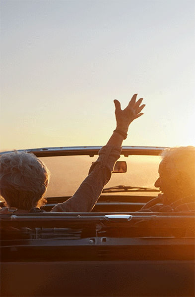 Zwei Personen sitzen in einem roten Cabrio mit offenem Verdeck und erhobenen Armen und blicken auf einen Sonnenuntergang über einer Berglandschaft. 