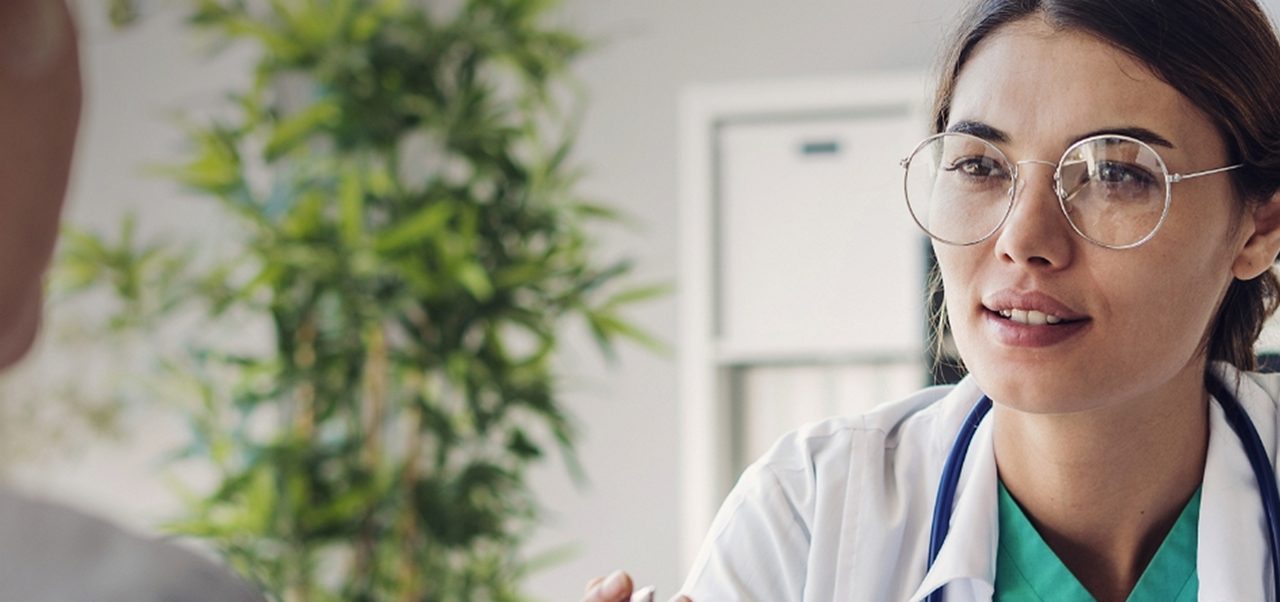 Ein Arzt, der eine Brille und einen weißen Kittel trägt, hört einem Patienten während einer Sprechstunde in einer Arztpraxis zu.