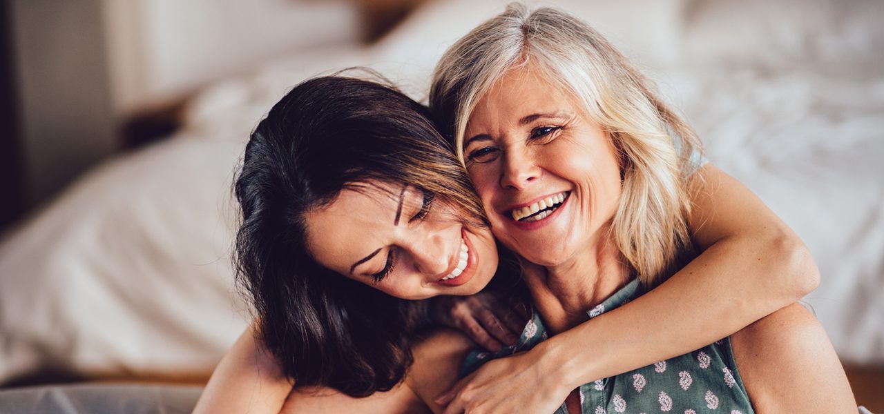 Zwei Frauen sitzen eng beieinander, lächeln und umarmen sich. Eine Frau hat den Arm um die andere gelegt, und sie scheinen einen warmen Moment im Haus vor einem unscharfen Hintergrund zu genießen.