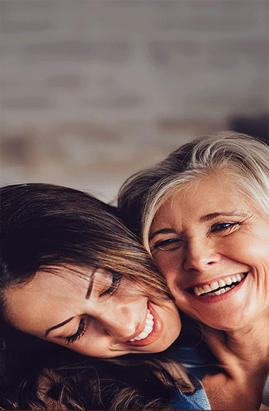 Zwei Frauen sitzen eng beieinander, lächeln und umarmen sich. Eine Frau hat den Arm um die andere gelegt, und sie scheinen einen warmen Moment im Haus vor einem unscharfen Hintergrund zu genießen. 