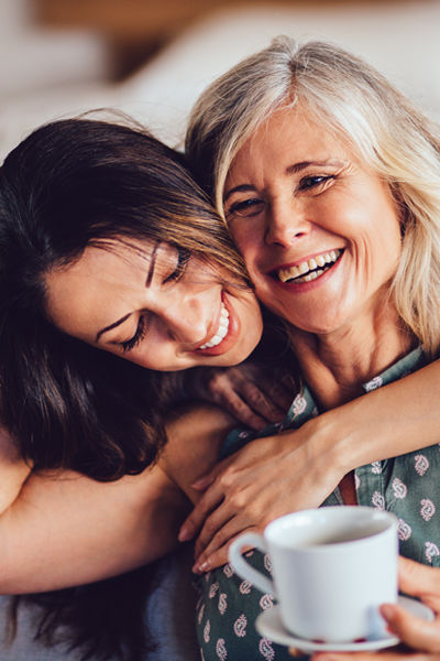 Zwei Frauen sitzen eng beieinander, lächeln und umarmen sich. Eine Frau hat den Arm um die andere gelegt, und sie scheinen einen warmen Moment im Haus vor einem unscharfen Hintergrund zu genießen. 