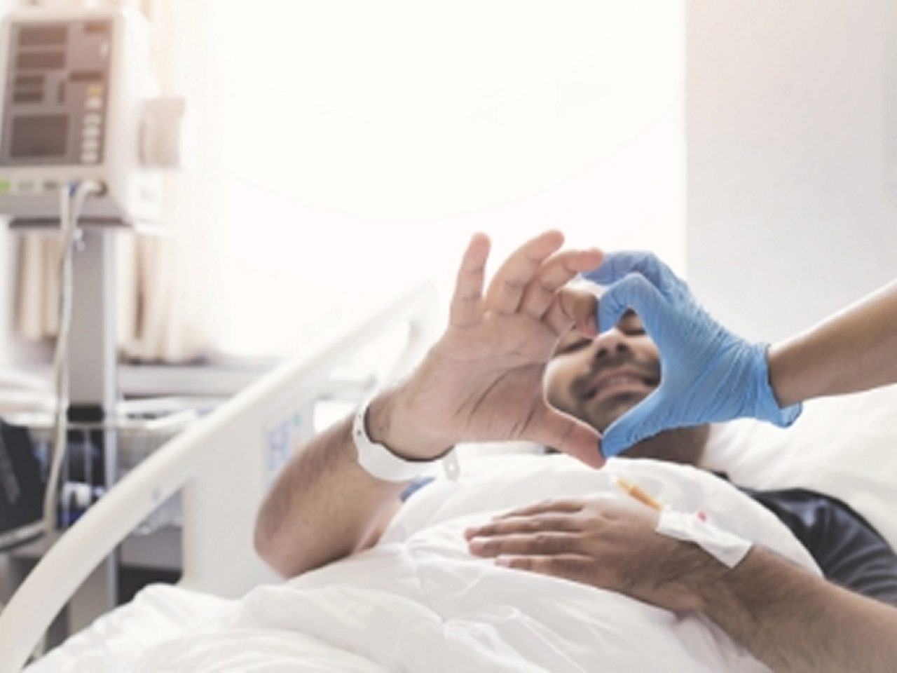 Ein Patient in einem Krankenhausbett formt mit seiner Hand ein Herz und verbindet seine Finger mit denen einer behandschuhten Pflegekraft.
