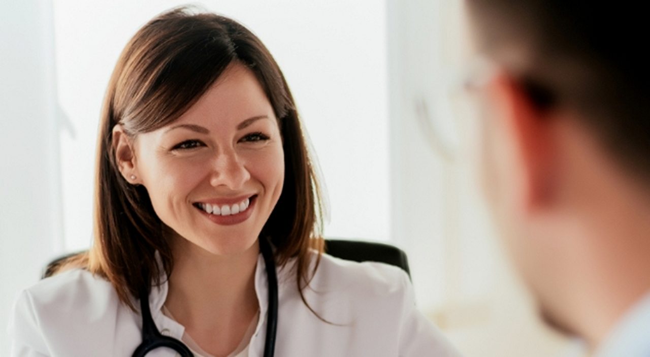 Eine Ärztin mit einem Stethoskop lächelt, während sie in einer Praxis mit einem Patienten spricht.