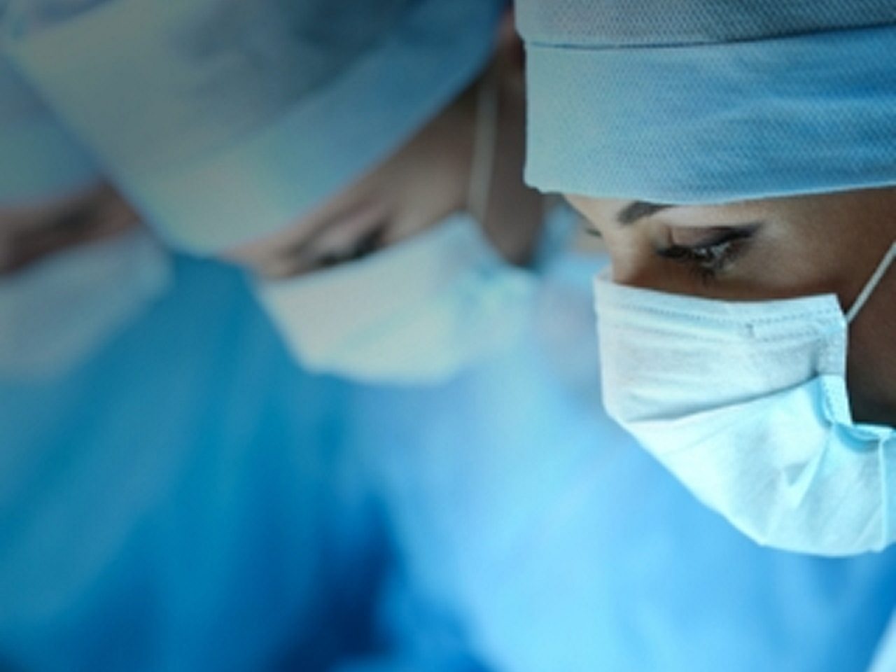 Drei Chirurgen in blauen Kitteln, Masken und Mützen konzentrieren sich während eines medizinischen Eingriffs in einem Operationssaal.