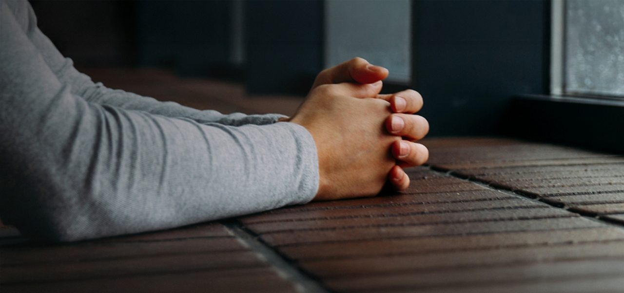 Eine Person in einem grauen langärmeligen Hemd sitzt mit gefalteten Händen auf einer gemauerten Fensterbank und schaut an einem regnerischen Tag aus dem Fenster.