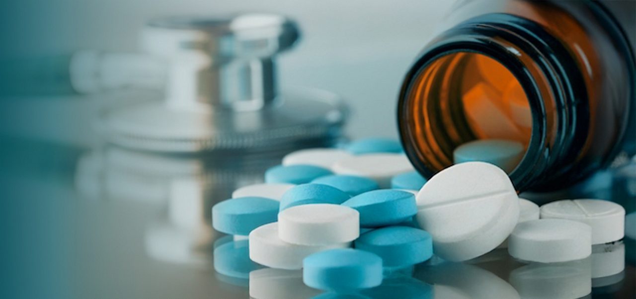 Ein braunes Pillenfläschchen liegt auf der Seite, aus dem blaue und weiße Tabletten herausquellen; ein Stethoskop ist unscharf im Hintergrund zu sehen.