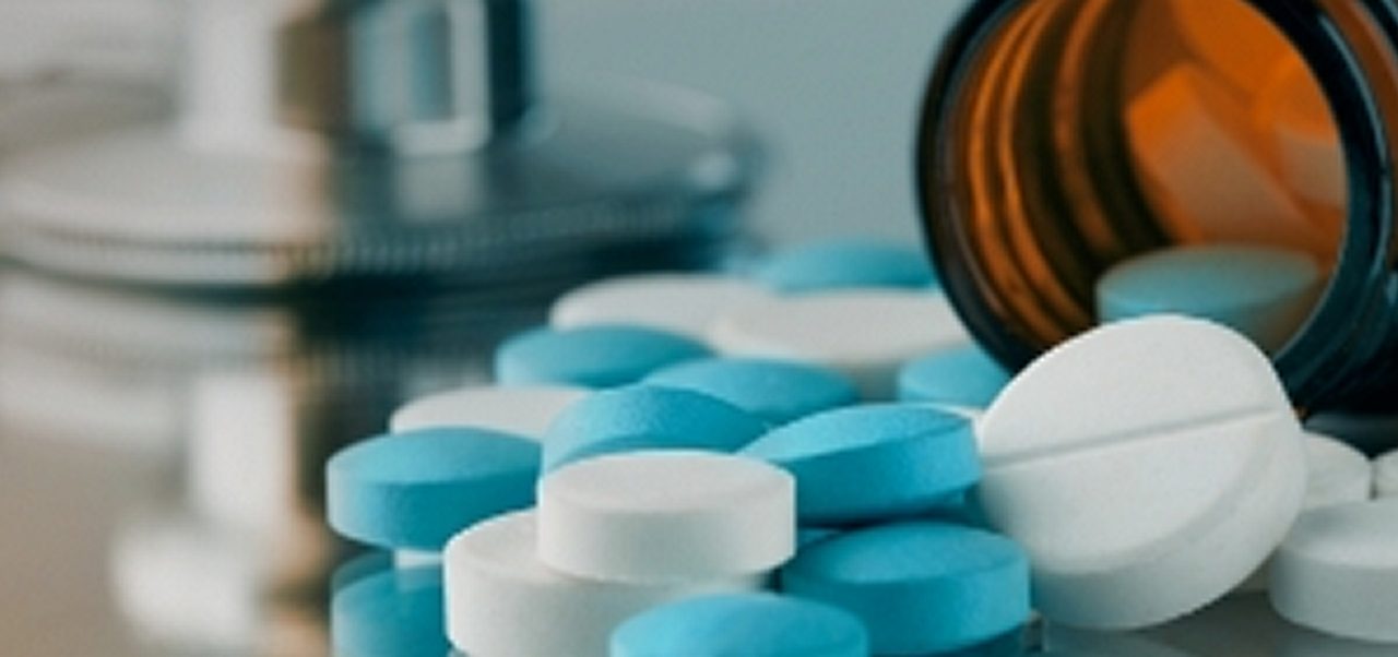 Ein braunes Pillenfläschchen liegt auf der Seite, aus dem blaue und weiße Tabletten herausquellen; ein Stethoskop ist unscharf im Hintergrund zu sehen.