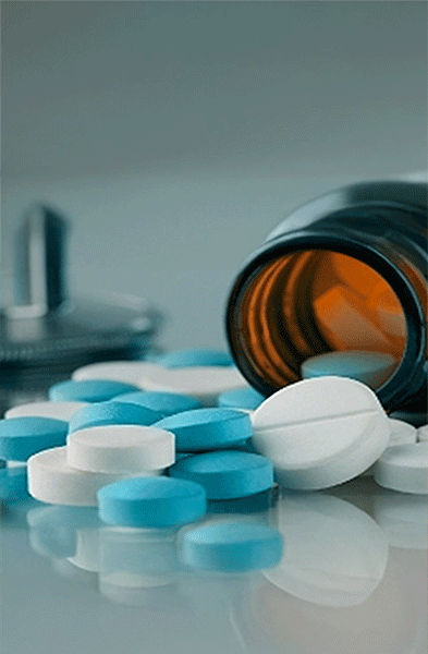 Ein braunes Pillenfläschchen liegt auf der Seite, aus dem blaue und weiße Tabletten herausquellen; ein Stethoskop ist unscharf im Hintergrund zu sehen.