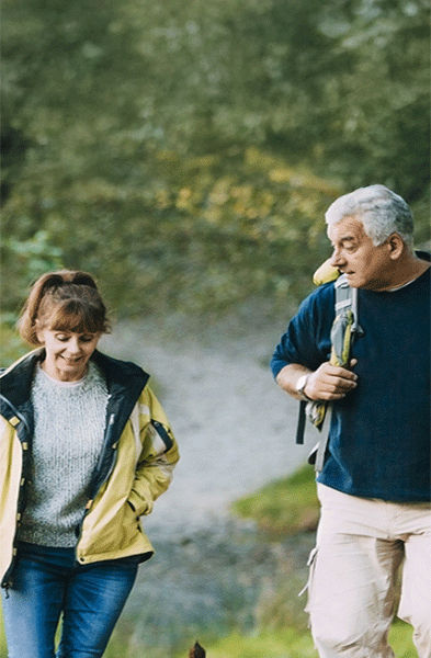 Zwei Erwachsene und ein Hund gehen bei Tageslicht gemeinsam auf einem felsigen Pfad durch ein Waldgebiet.
