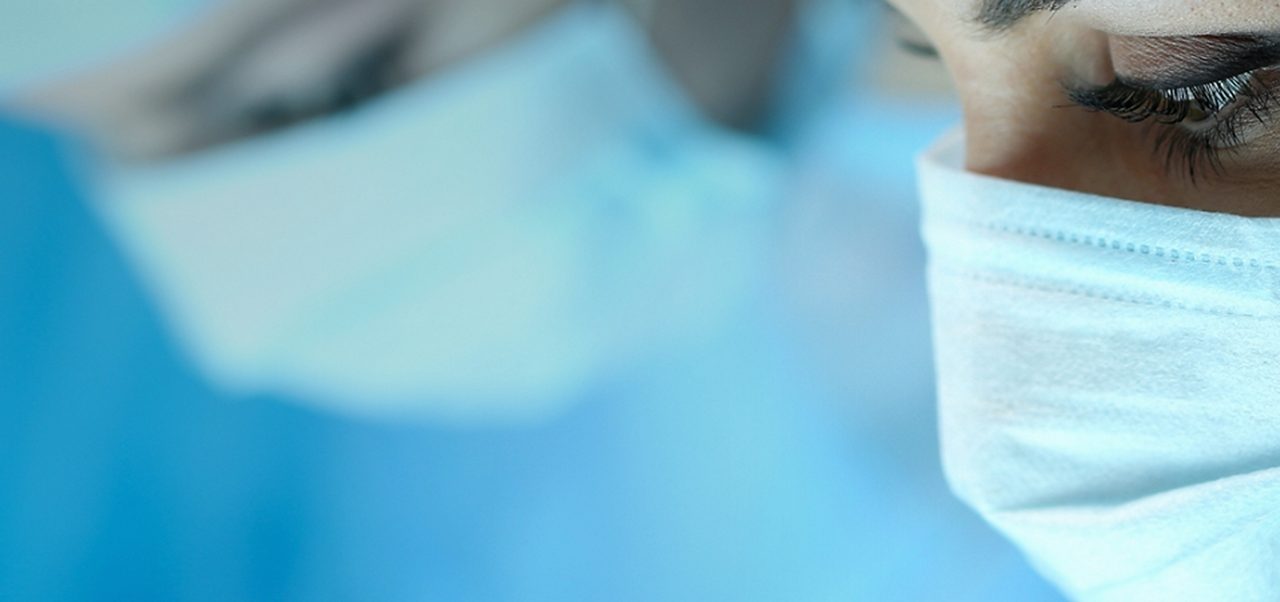 Drei Chirurgen in blauen Kitteln, Masken und Mützen konzentrieren sich während eines medizinischen Eingriffs in einem Operationssaal.