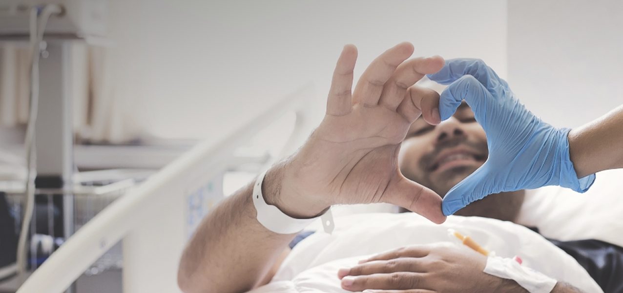Ein Patient in einem Krankenhausbett formt mit seiner Hand ein Herz und verbindet seine Finger mit denen einer behandschuhten Pflegekraft.