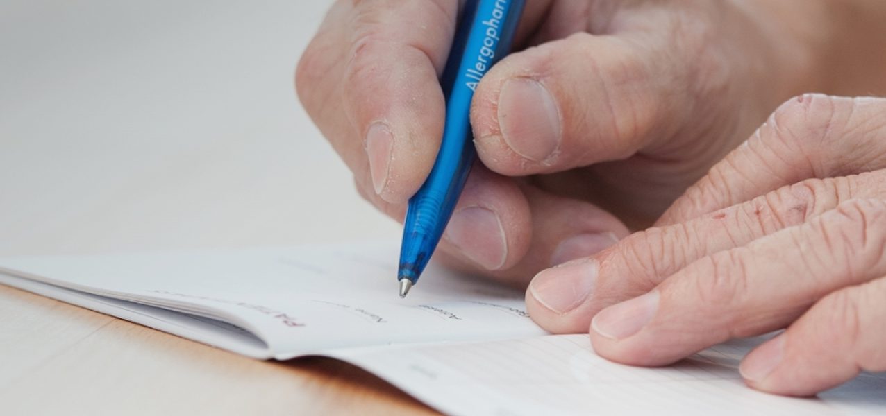 Eine Person, die einen Ehering trägt, schreibt mit einem blauen Stift auf ein Dokument, während sie ihre Hand auf einen Holztisch stützt.