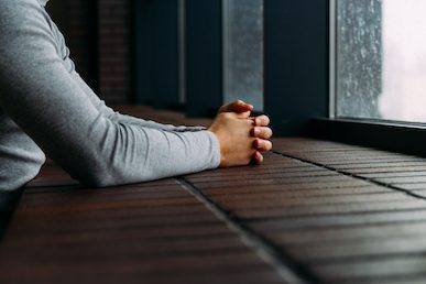 Eine Person mit gefalteten Händen stützt ihre Arme auf eine hölzerne Oberfläche und schaut an einem regnerischen Tag aus dem Fenster.