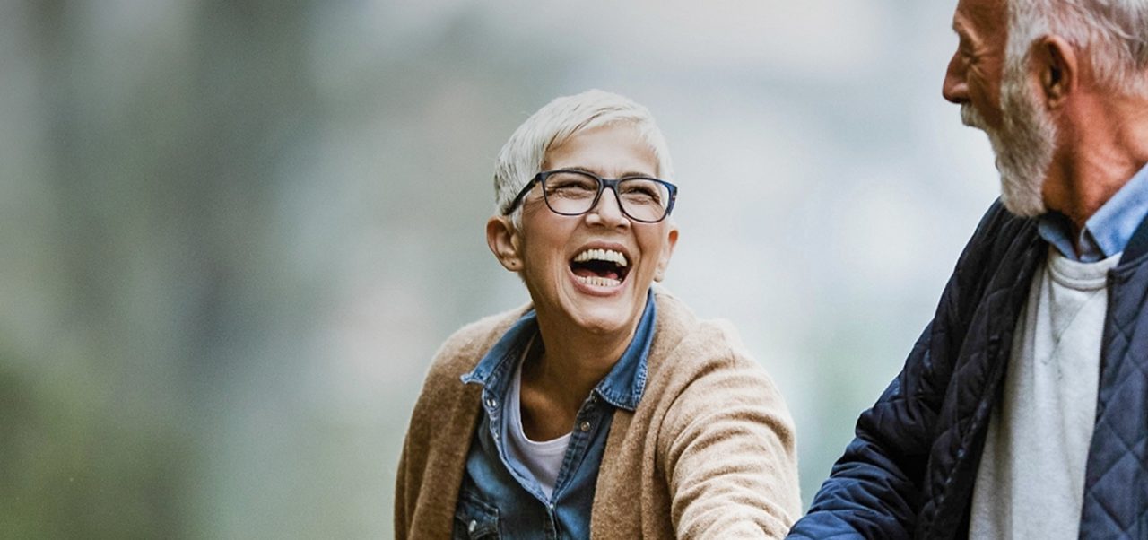 Ein älterer Mann und eine ältere Frau gehen im Freien spazieren, halten sich an den Händen, lächeln und lachen, tragen legere Jacken und sind von Grün umgeben.
