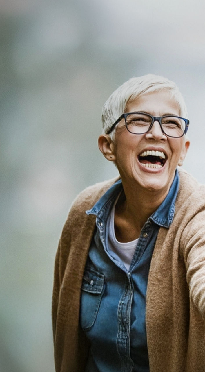 Ein älterer Mann und eine ältere Frau gehen im Freien spazieren, halten sich an den Händen, lächeln und lachen, tragen legere Jacken und sind von Grün umgeben.