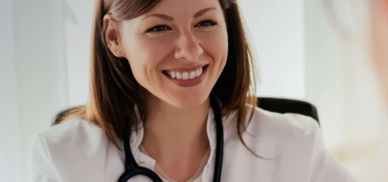 Eine lächelnde Ärztin mit einem Stethoskop spricht mit einem männlichen Patienten in einem hellen Büro.