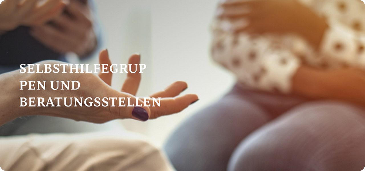 Drei Personen sitzen in einer Diskussion; eine Person hält ein Klemmbrett, eine andere gestikuliert mit den Händen und die dritte sitzt mit gefalteten Händen.
