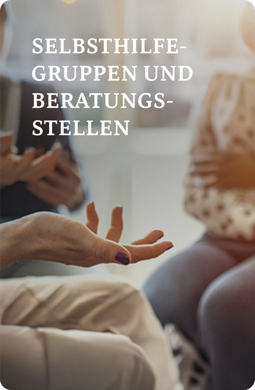 Drei Personen sitzen in einer Diskussion; eine Person hält ein Klemmbrett, eine andere gestikuliert mit den Händen und die dritte sitzt mit gefalteten Händen.