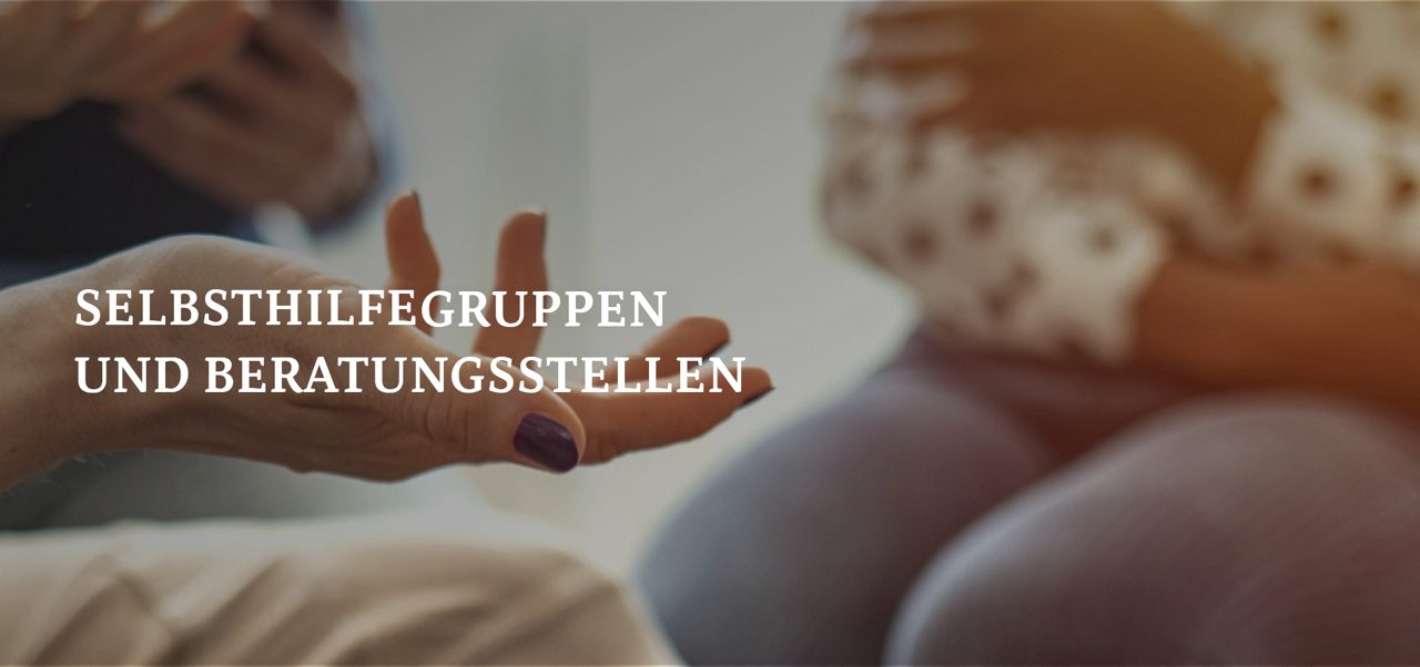 Drei Personen sitzen in einer Diskussion; eine Person hält ein Klemmbrett, eine andere gestikuliert mit den Händen und die dritte sitzt mit gefalteten Händen.